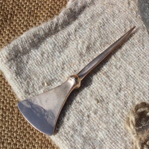 May include: A silver-coloured metal tool with a pointed end and a flat, angled blade. It rests on a light grey, textured fabric. The background includes a woven, tan fabric and a bundle of twine.