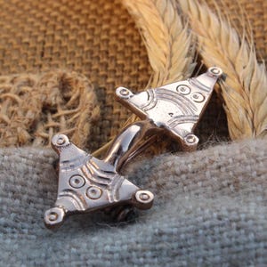 May include: A silver-toned metal brooch with a stylized design. The brooch features two triangular elements connected by a curved bar. Each triangle has a pattern of circles and lines. The brooch is set against a textured background.