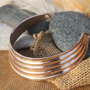 May include: A copper-colored cuff bracelet with three horizontal ridges and a decorative edge. The bracelet is open at one end. The background includes gray stones, burlap, and a textured fabric.
