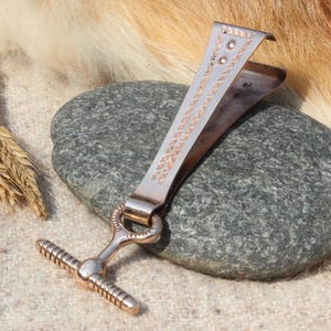 May include: A bronze-coloured metal tie clip with a decorative design, resting on a grey stone. The clip has a T-bar and a detailed pattern. Two stalks of wheat are to the left.