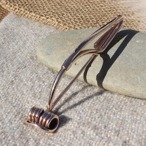 May include: A copper-colored hair barrette with a unique design. The barrette features a coiled base and a curved, openwork top. The barrette is resting on a light gray stone, with a textured fabric background.