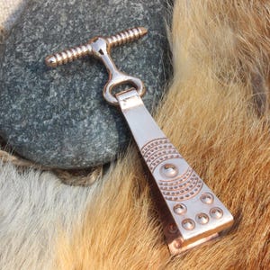 May include: A copper-coloured metal bottle opener with a T-shaped handle and an engraved design. The opener is on a grey stone and fur background, highlighting its intricate details and craftsmanship.