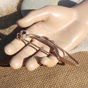 May include: A copper wire brooch in the shape of a bow and arrow. The brooch is handmade and has a rustic, organic look.