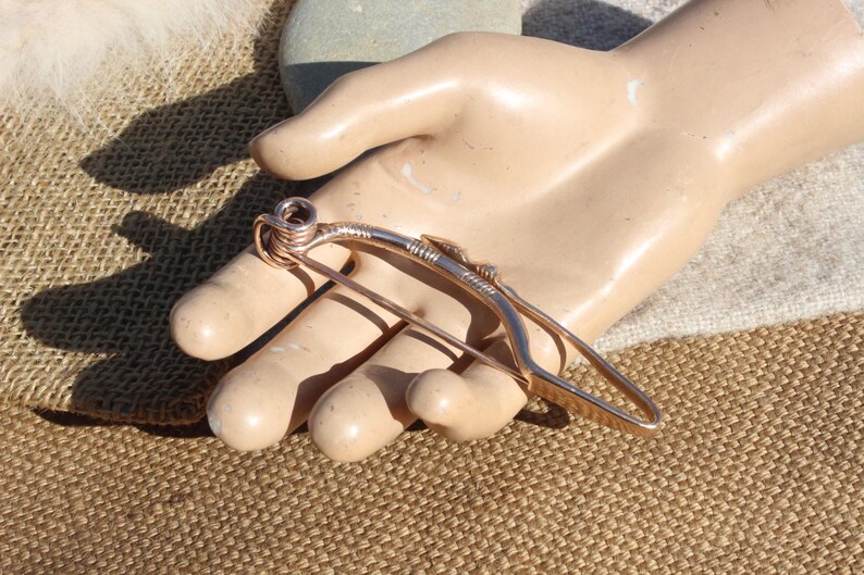May include: A copper wire brooch in the shape of a bow and arrow. The brooch is handmade and has a rustic, organic look.