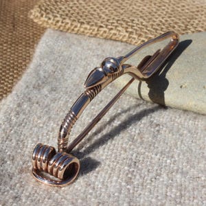 May include: A bronze brooch with a stylized bird design. The brooch has a coiled wire detail at the bottom and a pin on the back.