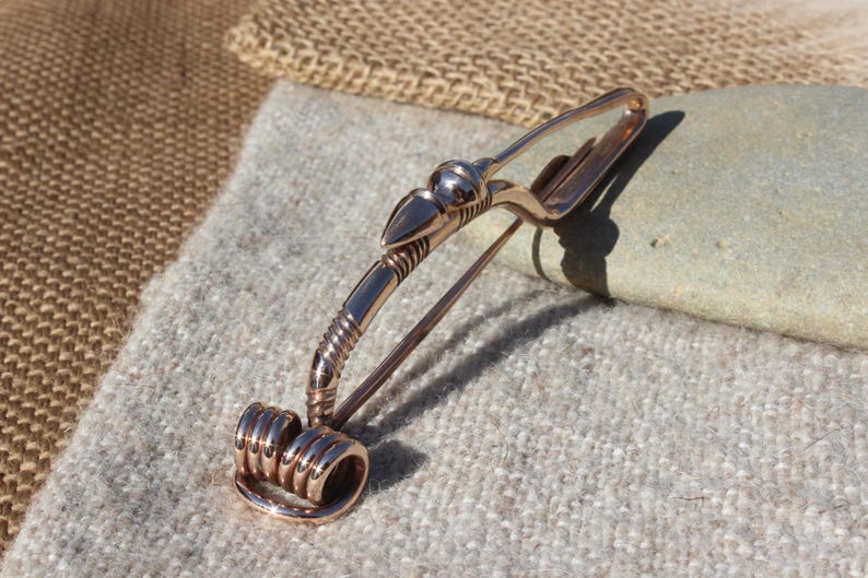 May include: A bronze brooch with a stylized bird design. The brooch has a coiled wire detail at the bottom and a pin on the back.