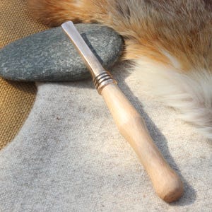 May include: A wooden-handled tool with a metal blade and a silver band around the handle. The tool is lying on a light grey fabric surface.