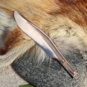 Könnte beinhalten: Eine metallische, blattförmige Klinge mit einer polierten, reflektierenden Oberfläche. Die Klinge hat eine spitz zulaufende Form und einen flachen Griffbereich mit zwei kleinen, hervorstehenden zylindrischen Stiften und einem Mittelgrat.