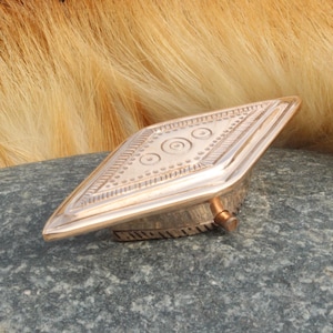 Op de afbeelding: Een ruitvormige koperen riemgesp met een reliëf geometrisch patroon van concentrische cirkels en lineaire randen. Het metalen oppervlak heeft een gepolijste afwerking en een kleine cilindrische pin is zichtbaar aan de zijkant van de gesp.