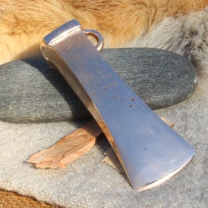 May include: A silver metal axe head with a curved blade, lying on a grey stone and a piece of wood. The axe head has a loop at the top. The background includes fur and a textured fabric.