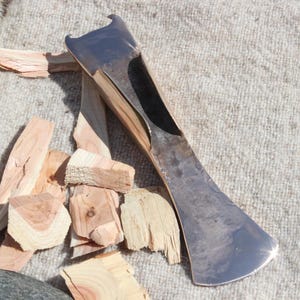 May include: A shiny metal axe head with a curved blade and a wooden handle, placed on a textured, light-coloured surface. There are pieces of light-coloured wood scattered around the axe head, suggesting a wood-splitting tool.