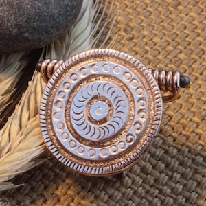May include: A bronze-colored, circular pendant with a spiral design and a pin. The pendant has a light blue center and is surrounded by a detailed border. The pin has a coiled design and a small, dark bead at the end. The pendant is on a woven surface.