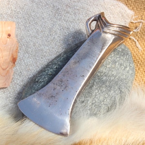 May include: A silver axe head with a curved blade sits on a grey stone. The axe has a handle with a loop at the top. Beside it are pieces of wood. The background includes textured fabric and fur.