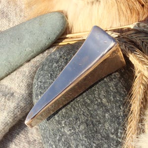 Peut inclure: Un pendentif triangulaire argenté et doré repose sur une pierre grise foncée, avec une pierre grise claire et des épis de blé en arrière-plan. Le pendentif a une surface polie et réfléchissante et un design géométrique.