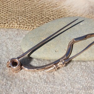 May include: A bronze Viking age brooch with a long pin and a decorative design. The brooch is resting on a gray rock.