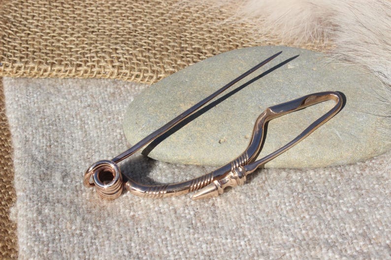 May include: A bronze Viking age brooch with a long pin and a decorative design. The brooch is resting on a gray rock.