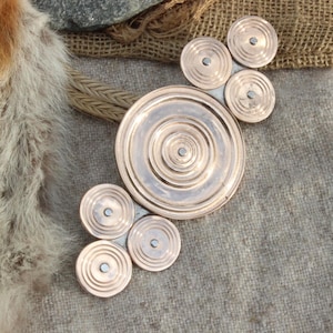 May include: A circular copper brooch with concentric circles and a smaller circle in the center. The brooch is made of multiple pieces that are joined together.
