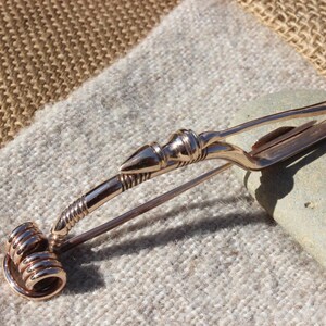 May include: A bronze Viking-style brooch with a spiral design and a pointed end. The brooch is on a brown and white fabric background.