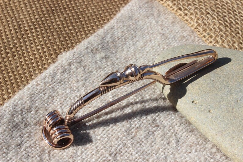 May include: A bronze Viking-style brooch with a spiral design and a pointed end. The brooch is on a brown and white fabric background.