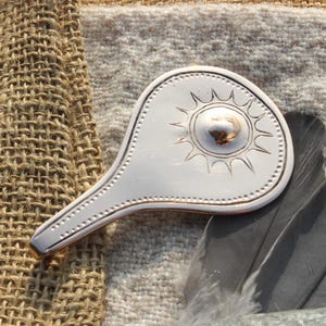 May include: A silver-colored, teardrop-shaped, decorative sun pendant with a raised, circular center and a sunburst design. The pendant is lying on a textured, brown fabric surface.