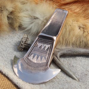 May include: A polished metal axe head featuring a wide, curved blade and a tapered handle section. The surface is decorated with intricate, hand-engraved geometric patterns, including rows of triangles and fine lines, creating a textured design.