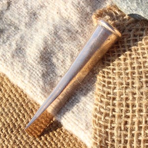 May include: A silver-colored, tapered metal object, possibly a tool or decorative item, lies on a textured surface of burlap and off-white fabric. The object has a cylindrical shape with a flat, angled end.