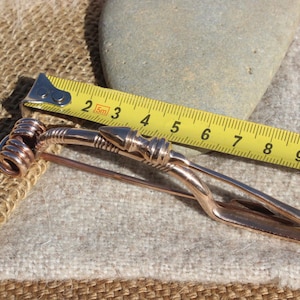 May include: A copper-colored metal brooch with a spiral design at one end and a pointed end at the other. The brooch is measured with a tape measure, showing it is approximately 10 inches long.