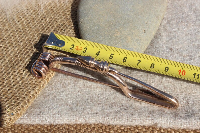 May include: A copper-colored metal brooch with a spiral design at one end and a pointed end at the other. The brooch is measured with a tape measure, showing it is approximately 10 inches long.