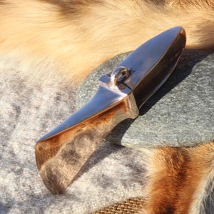 Op de afbeelding: Een gepolijst, zilverkleurig mes met een gebogen lemmet en een kleine ring. Het mes ligt op een grijze steen en een getextureerd, lichtgrijs oppervlak, met een achtergrond van bruine en tan vacht.