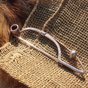 May include: A silver brooch with a coiled wire design at one end and a round ball at the other. The brooch is on a brown burlap background.