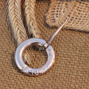May include: A vintage metal ring with a pointed extension, possibly a tool or jewelry component. The ring is a light metallic colour, with etched markings around its circumference. The background features wheat stalks and a textured brown fabric.