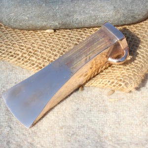 May include: A metal wedge tool with a polished silver blade and a bronze-coloured handle. The tool is lying on a burlap sack and a white cloth. A grey stone is in the background.