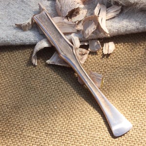 May include: A silver-colored, elongated tool with a curved handle and a flat, angled end, possibly a spatula or scraper. It rests on a textured, tan surface with wood shavings and a piece of light-colored fabric.