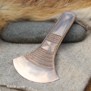May include: A polished copper-colored metal tool with a wide, curved blade and a narrow handle. The surface features an engraved pattern of repeating diagonal lines and a central diamond shape, creating a textured design on the metal surface.