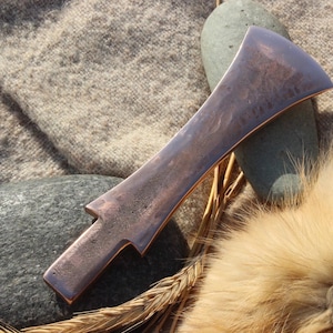 May include: A copper-coloured, hand-forged blade with a distinctive shape, resting on a stone and a textured fabric. The blade has a flat, elongated body with a curved edge and a notched handle. The background includes a stone, fur, and wheat.
