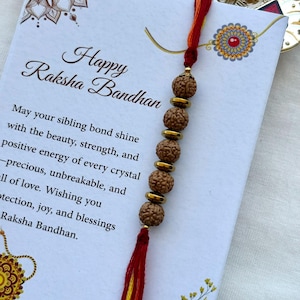 May include: A Raksha Bandhan greeting card with the text "Happy Raksha Bandhan" and a decorative bracelet. The bracelet features brown beads and gold-colored spacers, with a red and yellow tassel. The card has floral and mandala designs.