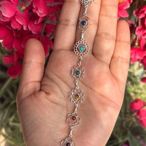 May include: A silver bracelet with seven chakra symbols, each with a different coloured gemstone. The bracelet is held against a backdrop of pink flowers. Gemstones include red, blue, green, yellow, and turquoise.