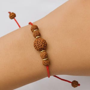 Peut inclure: Un bracelet fait à la main avec des perles marron texturées et des entretoises dorées enfilées sur un cordon rouge. Le bracelet est noué avec un nœud coulissant pour un ajustement réglable. Les perles ont un aspect organique unique.