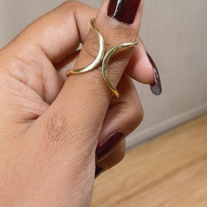 Könnte beinhalten: Ein goldfarbener Ring mit einem einzigartigen, offenen Design. Der Ring umschließt den Finger und erzeugt einen eleganten und modernen Look. Der Ring wird an einem Finger mit dunkelrotem Nagellack getragen.