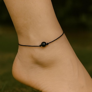May include: A black beaded anklet with a single, large, round bead at the center. The anklet is made of a thin, black cord and is worn on an ankle. The background is blurred green.