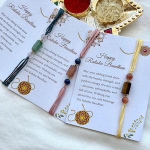May include: Three Raksha Bandhan bracelets on individual white cards with floral accents and the text Happy Raksha Bandhan. Each bracelet features coloured threads, small round beads, and a central rectangular gemstone in green, pink, or brown.