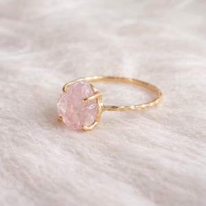 May include: A gold ring with a light pink, oval gemstone. The ring has a textured band and a simple design. The gemstone is held by four prongs. The ring's band has a hammered texture.
