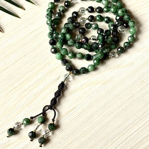 Puede incluir: Un mala de piedras preciosas verdes y negras con cuentas de cuarzo transparente y un borla negra. El mala está hecho de piedras naturales y está encordado en un cordón dorado.