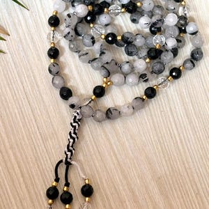 May include: A beaded necklace featuring a mix of black, white, and clear quartz beads. The necklace has a knotted tassel with black and white cord, and gold-colored accents. The beads are strung on a cord, creating a layered effect.