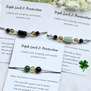 Può includere: Tre braccialetti con perline nere, gialle e verdi sono esposti su biglietti bianchi. I biglietti riportano "Triple Luck & Protection" ed elencano i cristalli utilizzati. I braccialetti sono infilati su un cordoncino grigio.