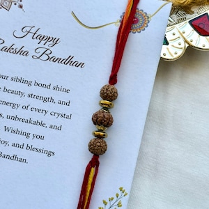 Puede incluir: Una pulsera Raksha Bandhan rakhi con tres cuentas marrones y espaciadores dorados, unida a hilos rojos y amarillos. La pulsera descansa sobre una tarjeta con el texto "Happy Raksha Bandhan" y elementos florales decorativos.
