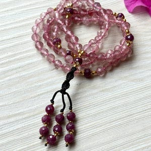 May include: A pink and clear crystal mala necklace with a tassel made of dark red beads. The necklace is made of faceted beads and has gold accents.