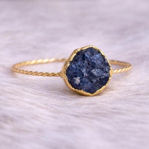 May include: A gold-colored ring featuring a rough-cut, deep blue gemstone. The gemstone is set in a gold bezel, and the band has a twisted rope design. The ring is displayed on a soft, white surface.