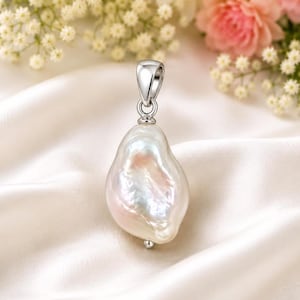 Puede incluir: Un colgante de perla barroca irregular con una fianza plateada. La perla muestra tonos iridiscentes de blanco, rosa y melocotón. El colgante se encuentra sobre un fondo de tela blanca y flores, sugiriendo un tema de joyería.