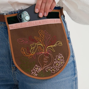 May include: A brown canvas waist pocket featuring a colorful embroidered beet and floral design. The pocket has a light pink velvet trim along the top edge, a dark green velvet inner lining, and a brown leather-like border, worn over blue denim jeans.